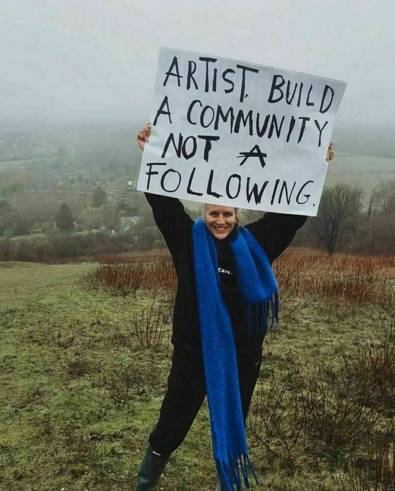 Author Amie McNee holds a large hand lettered sign that says “Artist, build a community not a following.” She is smiling dressed in cozy black sweats with a royal blue scarf draped around her neck. Beyond Amie you see the English countryside blanketed in fog.